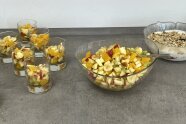 Eine große Schüssel und Portionsgläser mit Obstsalat. Im Hintergrund eine Schale mit Quarkspeise.