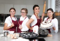 Vier Frauen in Küchenschürzen und mit Küchenutensilien und einer Torte.