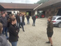 Im Hof eines landwirtschaftlichen Betriebs stehen junge Leute und sprechen mit einer FRau rechts im Bild.