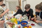 Kinder stehen um einen Tisch mit Schüsseln voller Weintrauben, Käsestücke, Tomaten, Gurken und Paprikastücke und bereiten damit bunte Spieße zu. 