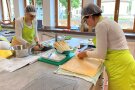 An zwei gegenüberliegenden Arbeitsplätzen in der Schulküche schält eine Frau Spargel und die andere trennt Backpapier von einer Platte Biskuitteig.