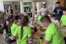 Kinder und Erwachsene sitzen um einen reich gedeckten Frühstückstisch mit Selbstgemachtem.