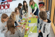 Acht Schülerinnen und eine Erwachsene stehen um einen Tisch mit Tellern voller Brotwürfeln, von denen sie sich eine Kostprobe nehmen.