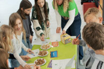 Acht Schülerinnen und eine Erwachsene stehen um einen Tisch mit Tellern voller Brotwürfeln, von denen sie sich eine Kostprobe nehmen.