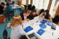 Schülerinnen und Schüler sitzen an quadratischen Tischen, auf denen allerlei Material, zum Beispiel blaue Umschläge liegen.