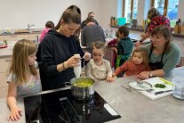 Kinder und Erwachsene stehen an enem herd. Eine Frau püriert eine Suppe.