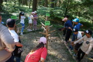 Eine Gruppe Kinder steht im Wald um drei kahle Stämme mit der Beschriftung Ahorn, Buche, Eiche. Mit Angeln versuchen sie, Plättchen mit abgebildeten Blättern an den zugehörigen Bäumen einzuhängen.