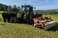 Männer und Frauen stehen in einer gemähten Wiese vor einem Schlepper mit Mähwerk.