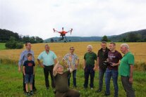 9 Personen stehen in Wiese und schauen auf Drohne über ihnen