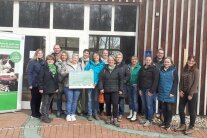 eine Gruppe Menschen vor einer Holz- und Fensterwand. Links davon ein Rollup 'Schule geht auf den Bauernhof'.