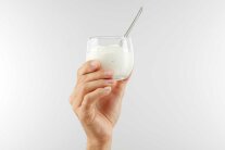Eine Hand hält rundliches Glas mit weißem Inhalt in die Höhe. Im Glas befindet sich Löffel.